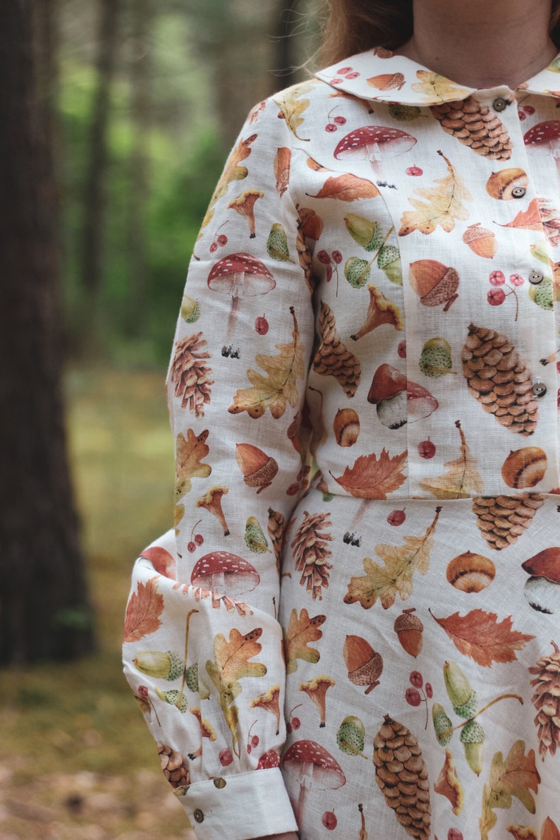 Puede incluir: Una camisa de lino blanca con cuello y mangas largas. La camisa presenta un estampado de hojas de oto&ntilde;o, bellotas, setas y pi&ntilde;as en tonos marrones, rojos y verdes.