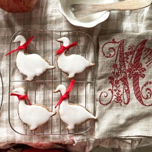 Puede incluir: Cuatro galletas blancas con forma de ganso decoradas con glaseado blanco y lazos de cinta roja en una rejilla de alambre plateado.