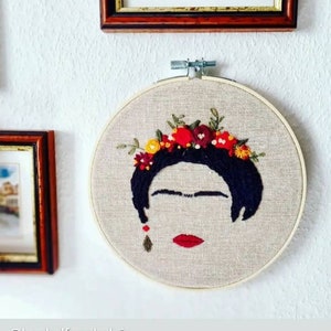 Op de afbeelding: Borduurwerk in een hoepel met een portret van Frida Kahlo met een bloemenkrans. Het portret is gemaakt met zwarte draad op een beige stoffen achtergrond. De bloemen zijn rood, geel en groen.