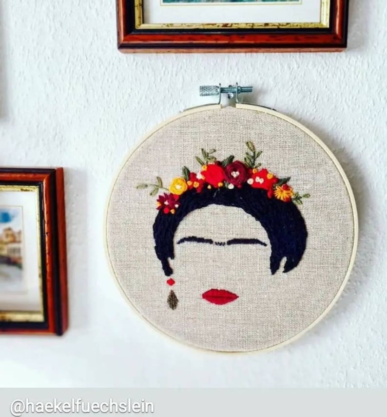 Op de afbeelding: Borduurwerk in een hoepel met een portret van Frida Kahlo met een bloemenkrans. Het portret is gemaakt met zwarte draad op een beige stoffen achtergrond. De bloemen zijn rood, geel en groen.