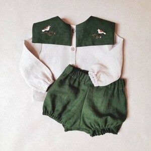 May include: A two-piece baby outfit featuring a light beige button-down shirt with a dark green collar embroidered with a bird and branch design. The outfit includes matching dark green bloomers.