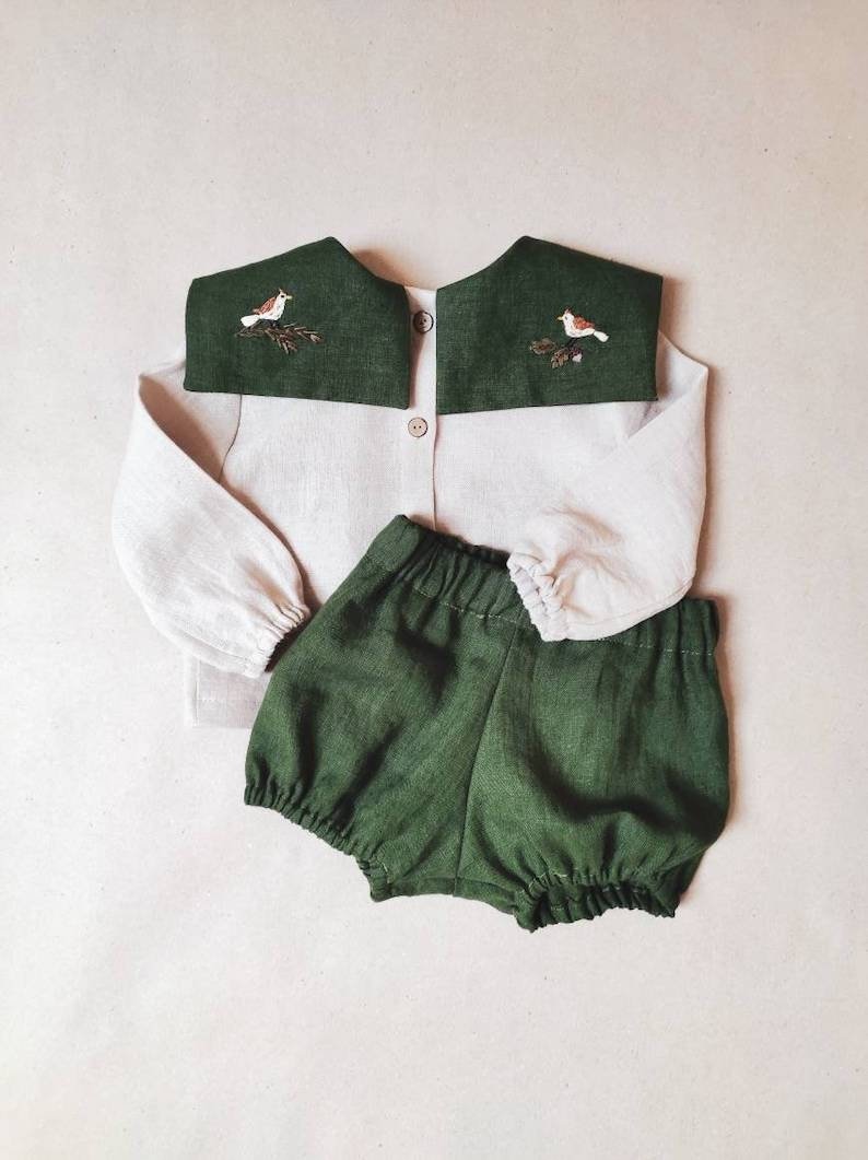 May include: A two-piece baby outfit featuring a light beige button-down shirt with a dark green collar embroidered with a bird and branch design. The outfit includes matching dark green bloomers.