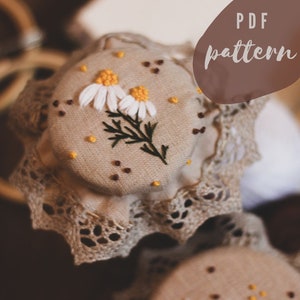 May include: A close-up of a beige fabric jar lid with a delicate lace trim. The lid is decorated with embroidered white daisies and green leaves, with a few brown and yellow dots scattered around. The text "PDF pattern" is visible in the upper right corner of the image.