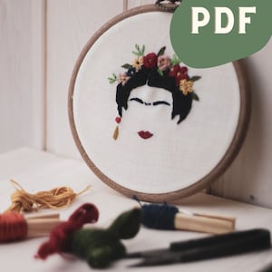 Op de afbeelding: Borduurring met een witte stoffen achtergrond waarop een geborduurd portret van een vrouw met een bloemenkrans te zien is. De vrouw heeft zwart haar, rode lippen en een gouden oorring. De ring is van hout en heeft een bruine afwerking. De afbeelding bevat ook borduurgaren in verschillende kleuren, een borduurschaar en een houten pin.