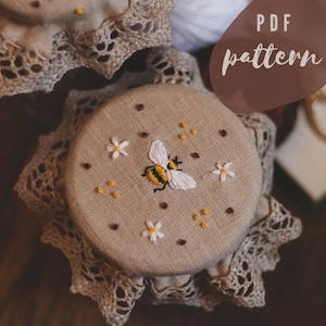 May include: A close-up of a beige fabric jar lid with a bee and daisy embroidery design. The lid is trimmed with a delicate lace and the text "PDF pattern" is visible in the background.