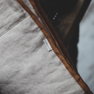 May include: Close-up of a linen pillow with a light beige center and a brown border. The pillow has a tag that reads "Ninis". The pillow is likely part of a home decor collection.