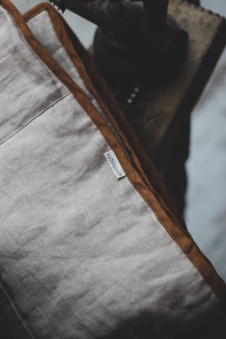 May include: Close-up of a linen pillow with a light beige center and a brown border. The pillow has a tag that reads "Ninis". The pillow is likely part of a home decor collection.