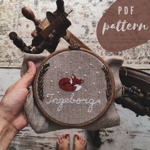 以下が含まれることがあります： 白い背景に赤いキツネが眠っている刺繍枠のクローズアップ。緑色のリースと「Ingeborg」という名前が白で刺繍されています。枠は人の手で持たれています。画像の右上に「PDFパターン」というテキストが書かれた茶色のラベルがあります。