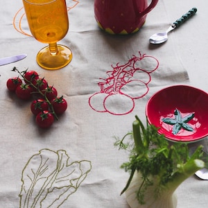 May include: A table setting featuring a red strawberry-shaped jug, a yellow glass, and a red bowl with a green star design. Fresh tomatoes and fennel are arranged on a linen tablecloth with embroidered vegetable designs.