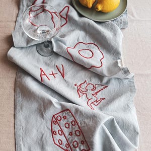 Op de afbeelding: Een lichtblauwe linnen tafelloper met rode geborduurde ontwerpen. De ontwerpen omvatten een glas, een ei, een hart met een pijl, een stuk kaas en de initialen "A+V".