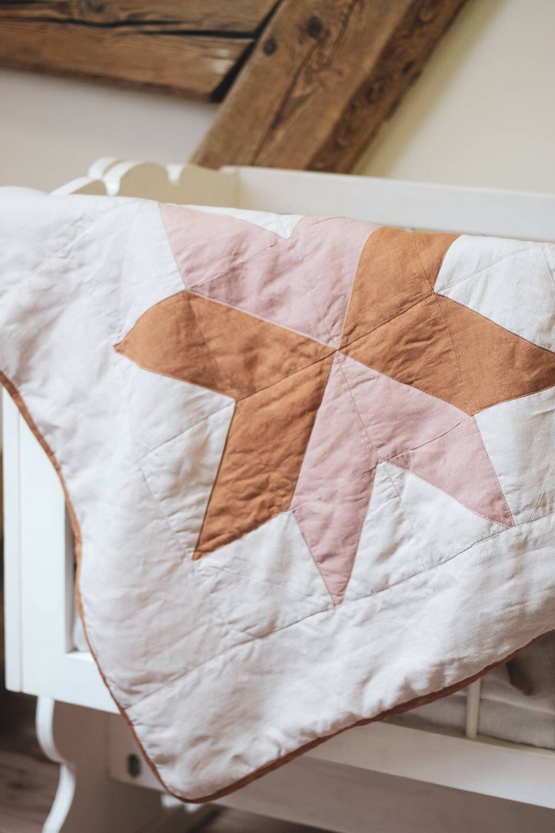 May include: A handmade baby quilt with a starburst design in shades of pink and brown on a white background. The quilt is placed over a white crib, showcasing its soft, quilted texture and decorative appeal. The quilt's edges are trimmed with a brown border.