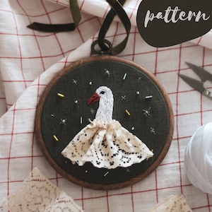 Pode incluir: Arte de bordado em aro com um pássaro branco com uma saia de renda, rodeado de glitter dourado e prateado sobre um fundo verde escuro. O aro é de madeira com um acabamento natural. O texto "PDF pattern" é visível no canto superior direito da imagem.