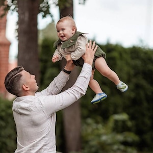 May include: A person holding a baby in the air. The baby is wearing a green and tan outfit with matching shoes. The person is wearing a light-colored button-down shirt. The background is a blurred green space.