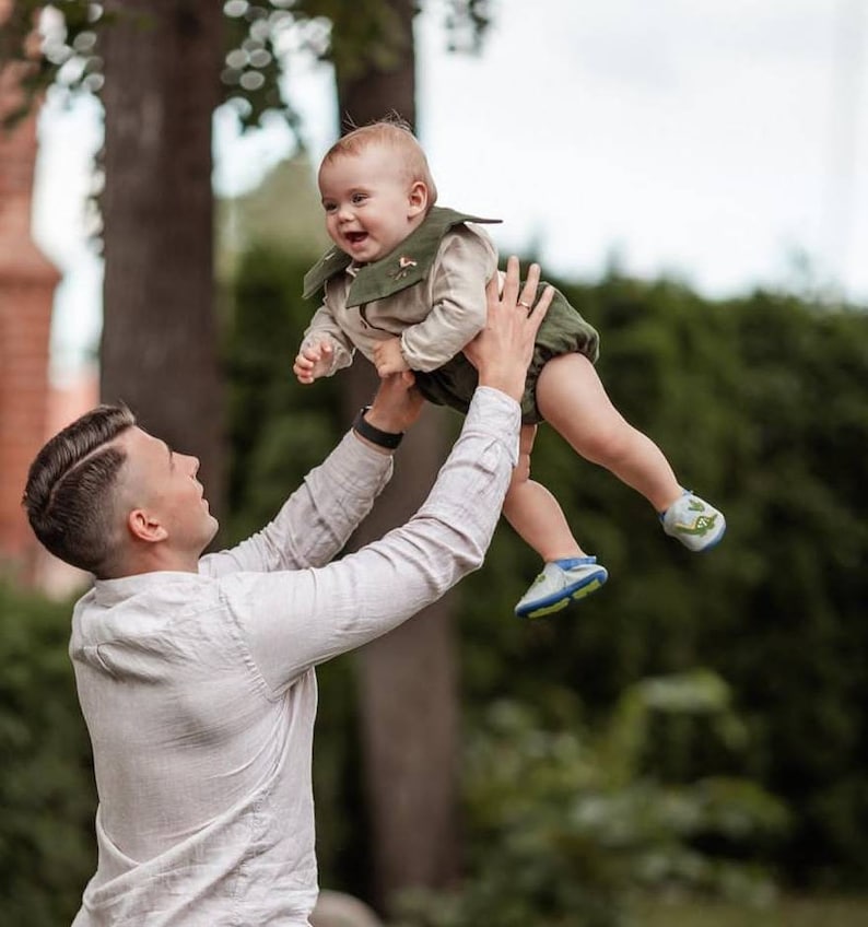 May include: A person holding a baby in the air. The baby is wearing a green and tan outfit with matching shoes. The person is wearing a light-colored button-down shirt. The background is a blurred green space.