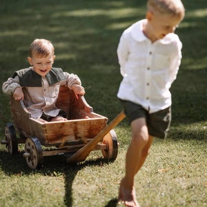 May include: A wooden toy wagon being pulled across a grassy lawn. A child is seated in the wagon, wearing a beige shirt with a dark green collar. Another child is pulling the wagon, wearing a white shirt and dark shorts.