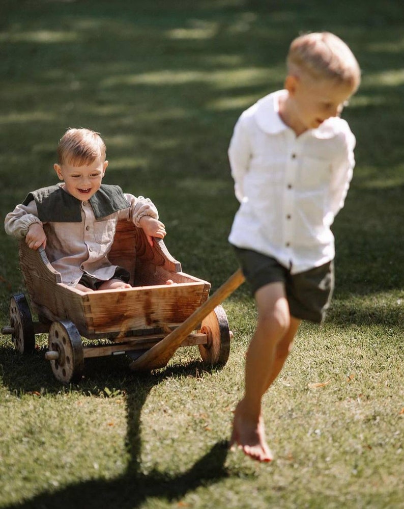 May include: A wooden toy wagon being pulled across a grassy lawn. A child is seated in the wagon, wearing a beige shirt with a dark green collar. Another child is pulling the wagon, wearing a white shirt and dark shorts.