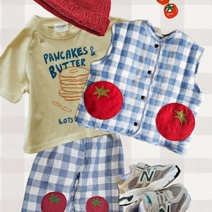 May include: A red knit beanie, a light yellow t-shirt with the text "PANCAKES & BUTTER LOTS", a blue and white gingham waistcoat and shorts with red tomato patches, and a pair of beige and grey trainers with the letter N on the side.