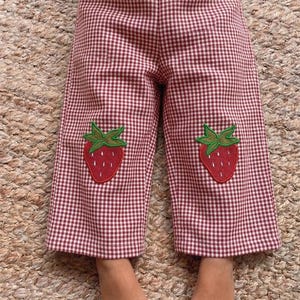 May include: Red and white gingham trousers featuring an elastic waistband and two embroidered strawberry patches, one on each leg. The trousers have a straight-leg cut and are crafted from a checkered fabric with a small-scale pattern throughout.