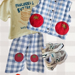 May include: A light yellow t-shirt with the text PANCAKES & BUTTER and a stack of pancakes, a blue and white gingham vest and matching pants with red tomato patches, and a pair of beige and grey athletic sneakers with a green N logo.