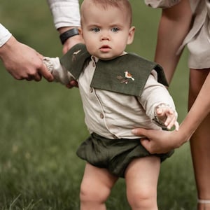 May include: A baby wearing a beige button-down shirt, dark green shorts, and a matching collar with an embroidered bird design. The baby is standing on green grass, with two adults holding the baby's arms.