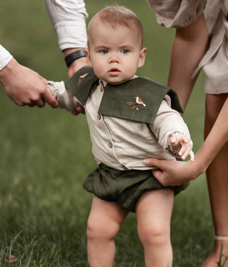 May include: A baby wearing a beige button-down shirt, dark green shorts, and a matching collar with an embroidered bird design. The baby is standing on green grass, with two adults holding the baby's arms.