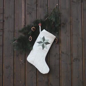Peut inclure: Une chaussette de Noël blanche avec une étoile verte et grise est suspendue à un mur en bois foncé, ornée d'une petite branche de conifère et d'ornements décoratifs. La chaussette est matelassée et possède une boucle pour l'accrocher.