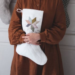 Peut inclure: Une chaussette blanche avec un motif de patchwork en forme d'étoile dans des tons de rose, vert et marron. La chaussette est tenue par une personne portant une robe marron avec un col blanc.