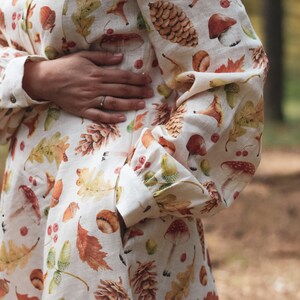 Puede incluir: Un vestido blanco de lino con estampado de oto&ntilde;o con hojas, bellotas, setas y pi&ntilde;as. El vestido tiene un cierre de botones en la parte delantera y mangas largas.