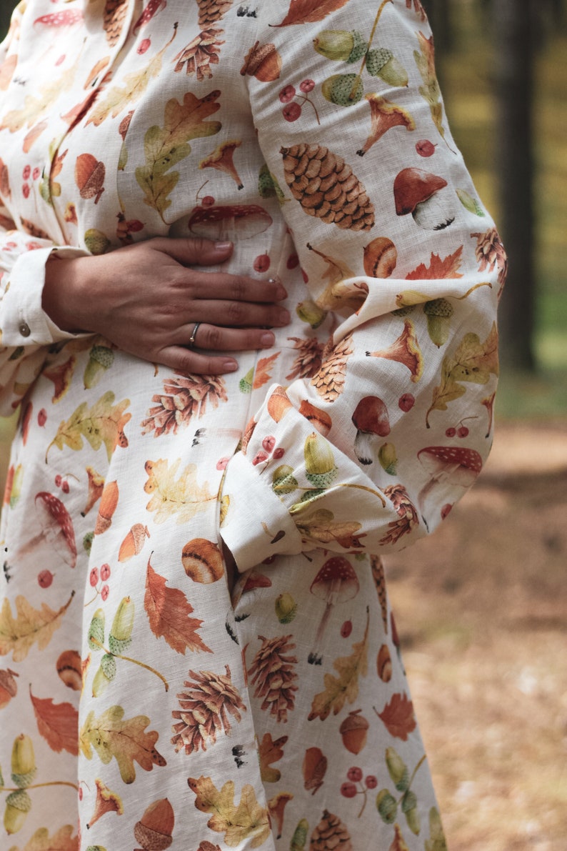 Puede incluir: Un vestido blanco de lino con estampado de oto&ntilde;o con hojas, bellotas, setas y pi&ntilde;as. El vestido tiene un cierre de botones en la parte delantera y mangas largas.