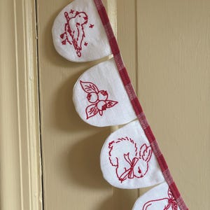 May include: A decorative banner with white fabric pennants, each embroidered with a different red design: a bird, berries, a rabbit, and pears. The pennants are attached to a red and white checkered ribbon.
