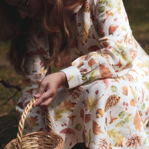 Puede incluir: Una mujer lleva un vestido de lino blanco con un estampado de oto&ntilde;o de hojas, bellotas y pi&ntilde;as y sostiene una cesta de mimbre.