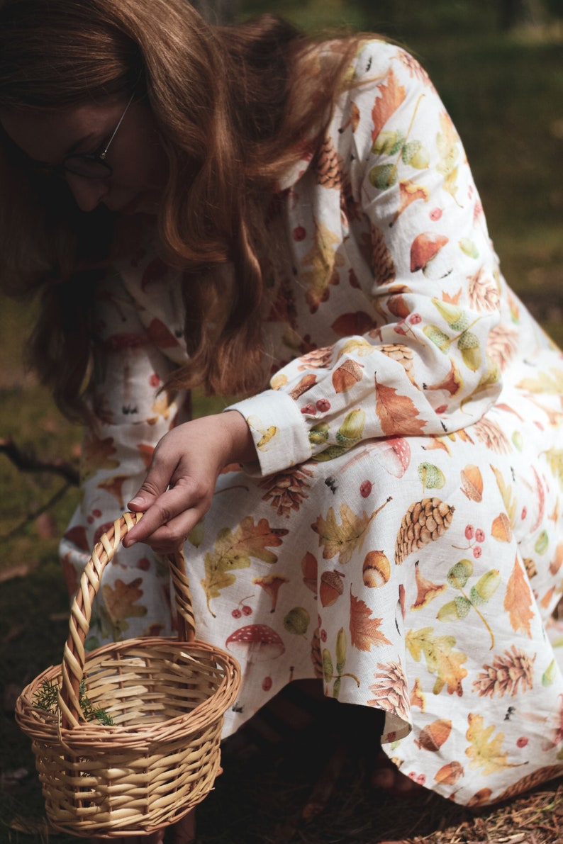 Puede incluir: Una mujer lleva un vestido de lino blanco con un estampado de oto&ntilde;o de hojas, bellotas y pi&ntilde;as y sostiene una cesta de mimbre.