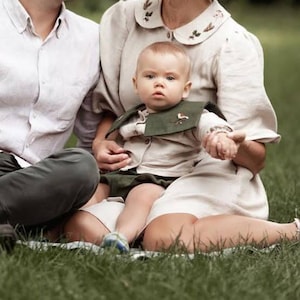 May include: A baby wearing a beige shirt and green shorts, with a matching green bib featuring a bird design. The baby is held by a person wearing a beige dress with floral embroidery on the collar, sitting on a green grassy surface.