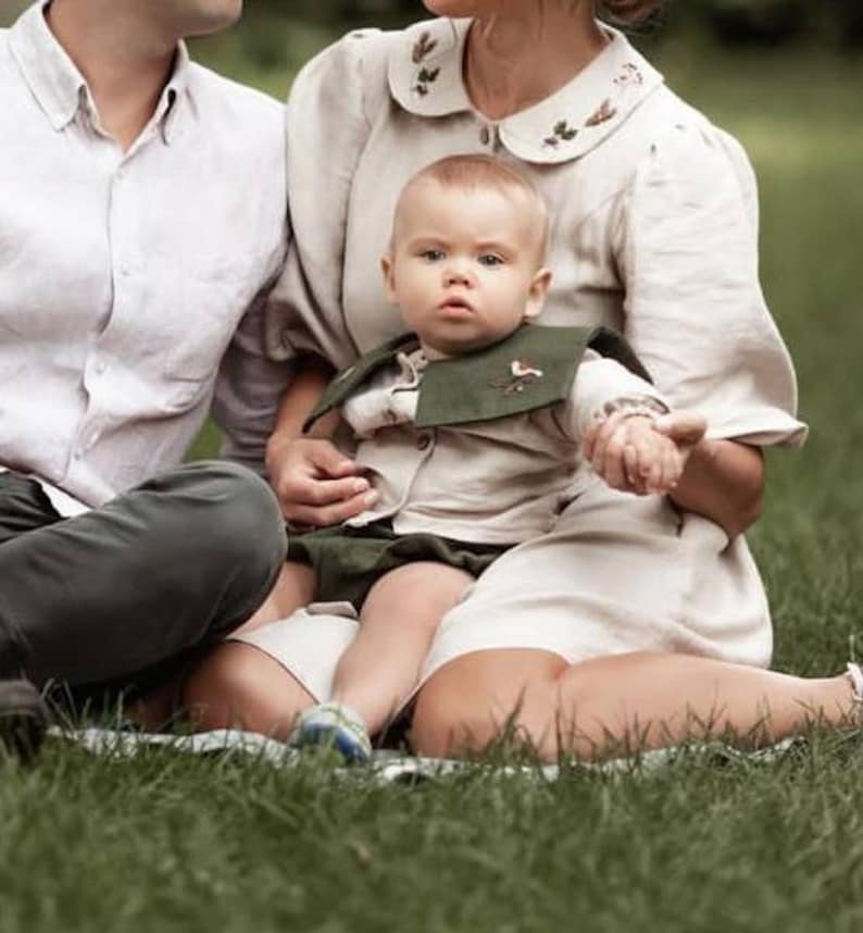 May include: A baby wearing a beige shirt and green shorts, with a matching green bib featuring a bird design. The baby is held by a person wearing a beige dress with floral embroidery on the collar, sitting on a green grassy surface.