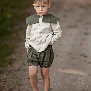 May include: A young child wearing a linen outfit. The outfit includes a light beige button-down shirt with a dark green collar and matching olive green bloomers. The child is also wearing brown sandals. The background is a blurred outdoor scene.