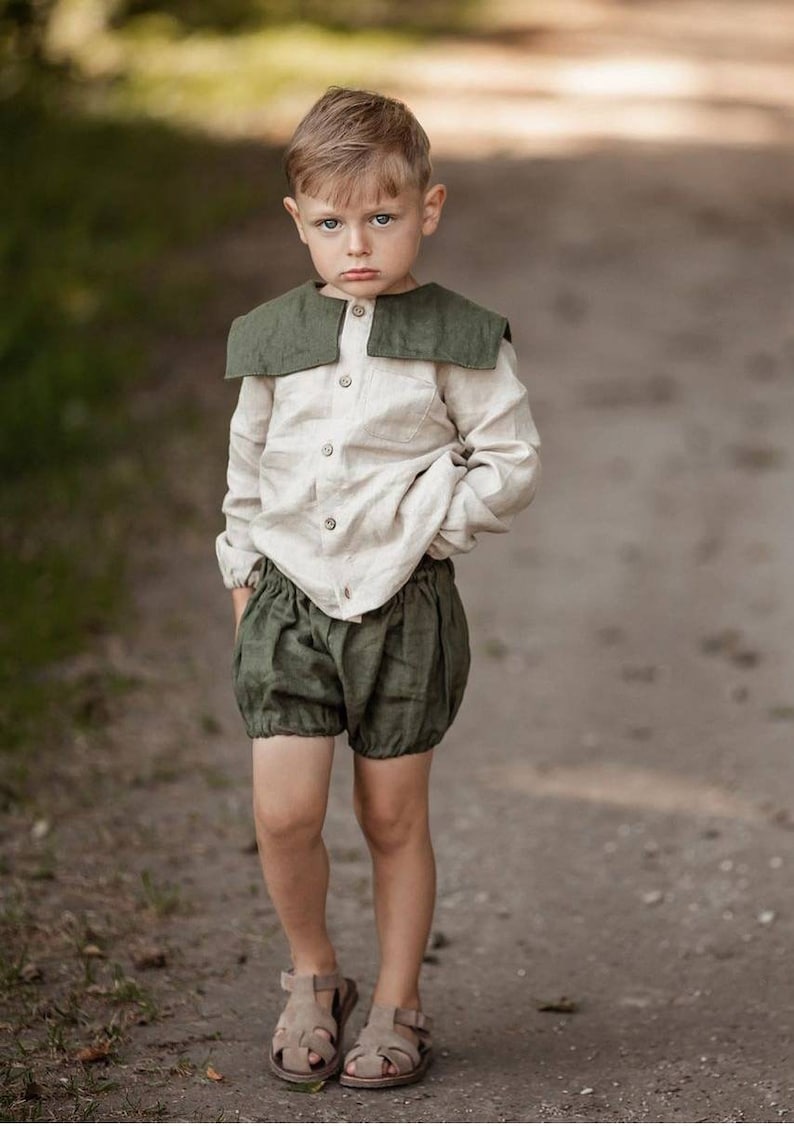 May include: A young child wearing a linen outfit. The outfit includes a light beige button-down shirt with a dark green collar and matching olive green bloomers. The child is also wearing brown sandals. The background is a blurred outdoor scene.