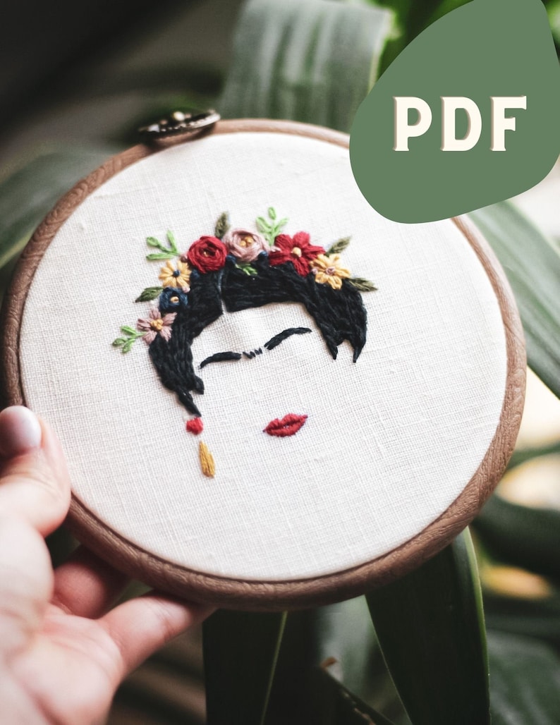 Op de afbeelding: Borduurwerk in een hoepel met een portret van een vrouw met een bloemenkrans. De vrouw heeft zwart haar, rode lippen en een gele oorbel. De hoepel is van hout en heeft een witte linnen achtergrond.