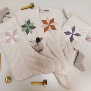 以下が含まれることがあります： 異なる色の星柄がついた、ベージュのキルティング加工が施されたクリスマスストッキングが4つあります。星は布製で、パッチワークのデザインになっています。ストッキングは、薄いベージュの表面に半円形に並べられています。