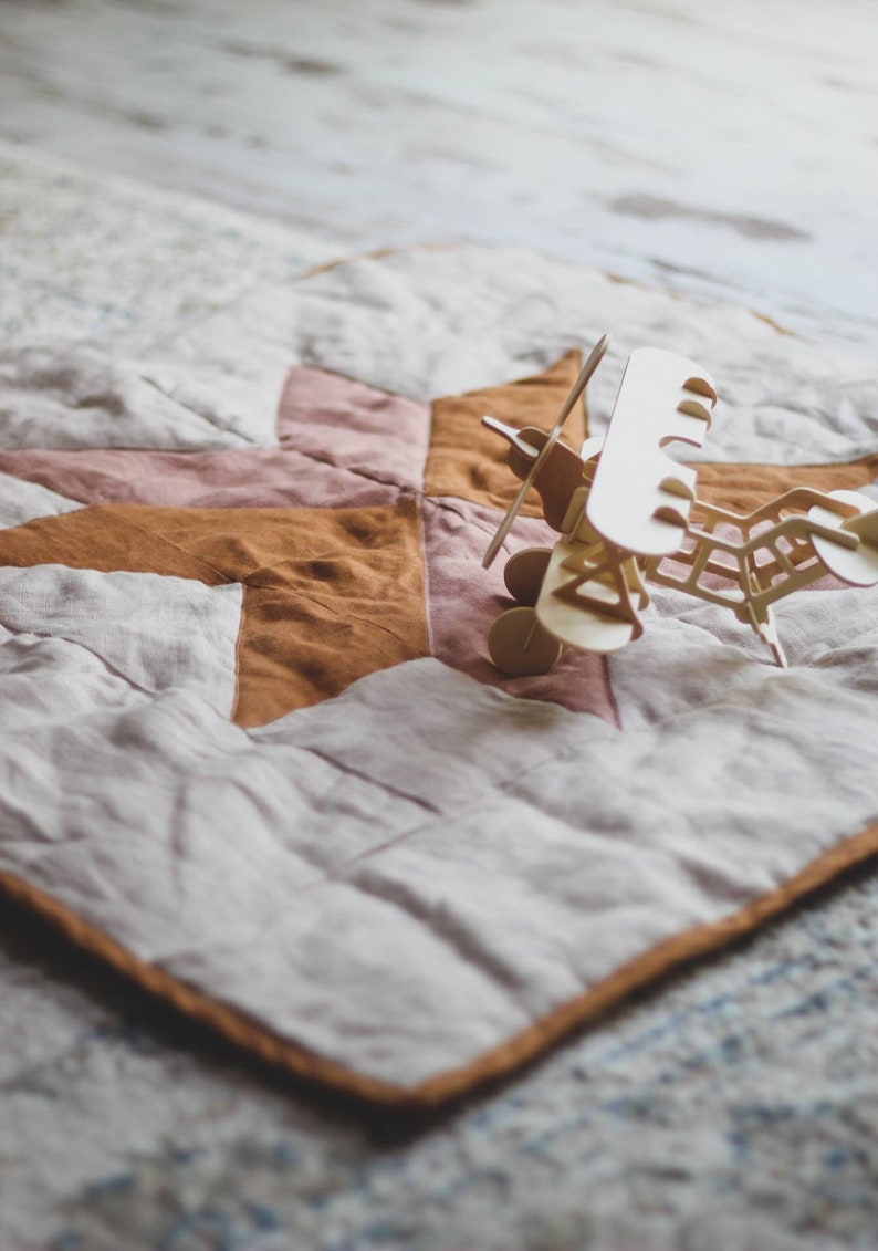 May include: A wooden toy airplane with a propeller and wheels sits on a star-patterned quilt. The quilt features a patchwork design in shades of pink, brown, and cream, with a brown border. The airplane is made of light-colored wood.