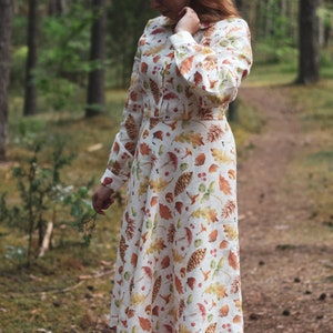 Puede incluir: Un vestido blanco de lino de manga larga con un estampado de oto&ntilde;o de hojas, bellotas y setas. El vestido tiene un cierre de botones en la parte delantera y una falda que llega a las rodillas.