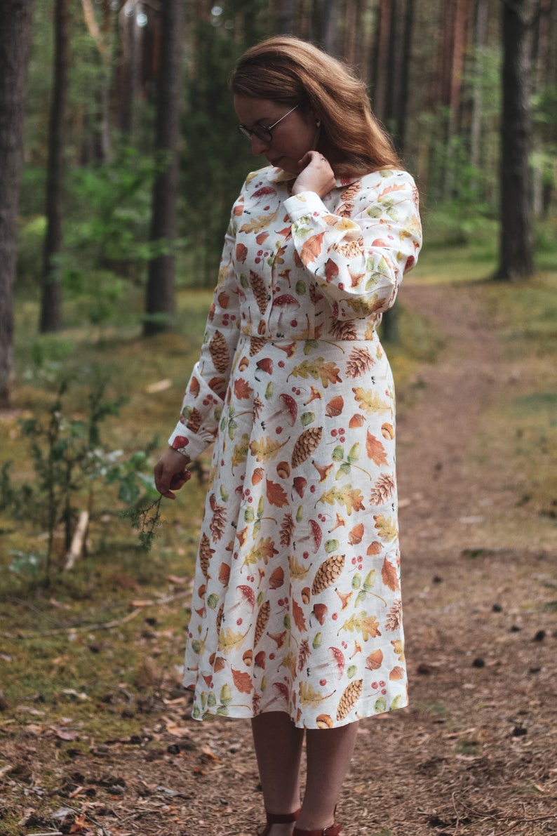 Puede incluir: Un vestido blanco de lino de manga larga con un estampado de oto&ntilde;o de hojas, bellotas y setas. El vestido tiene un cierre de botones en la parte delantera y una falda que llega a las rodillas.