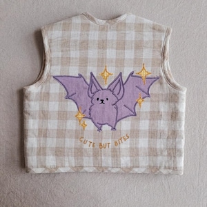 May include: A sleeveless waistcoat featuring a beige and white gingham pattern. The centre displays an embroidered purple bat with yellow star accents and the text "CUTE BUT BITES" stitched in yellow thread below the bat figure.