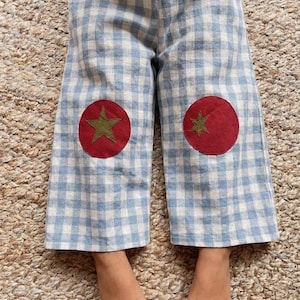 Op de afbeelding: Blauw-witte geruite kinderbroek met rode ronde patches met een groen sterontwerp op de knieën. De broek heeft een elastische tailleband en is gemaakt van een lichte stof.