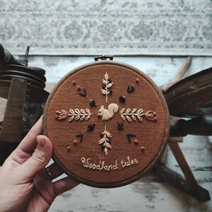May include: A round, brown embroidery hoop featuring a squirrel and floral design. The embroidery is in cream, brown, and black thread. The text "Woodland tales" is stitched at the bottom. The hoop is held by a hand.