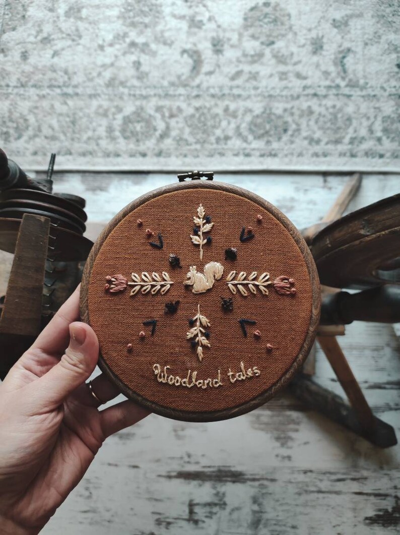 May include: A round, brown embroidery hoop featuring a squirrel and floral design. The embroidery is in cream, brown, and black thread. The text "Woodland tales" is stitched at the bottom. The hoop is held by a hand.