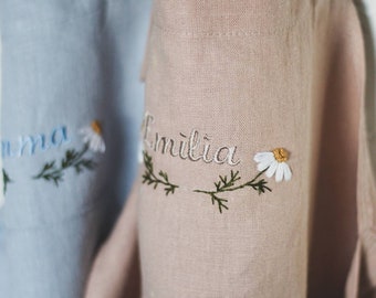 May include: Two linen aprons with embroidered names and flowers. The apron on the left is light blue with the name "Anna" embroidered in white. The apron on the right is light pink with the name "Emilia" embroidered in white.