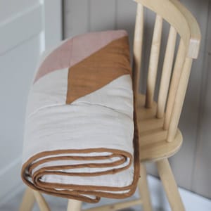 May include: A folded quilt with a geometric design in shades of white, pink, and brown rests on a light wooden chair. The quilt has a brown border and is neatly rolled. The chair is made of light-colored wood.