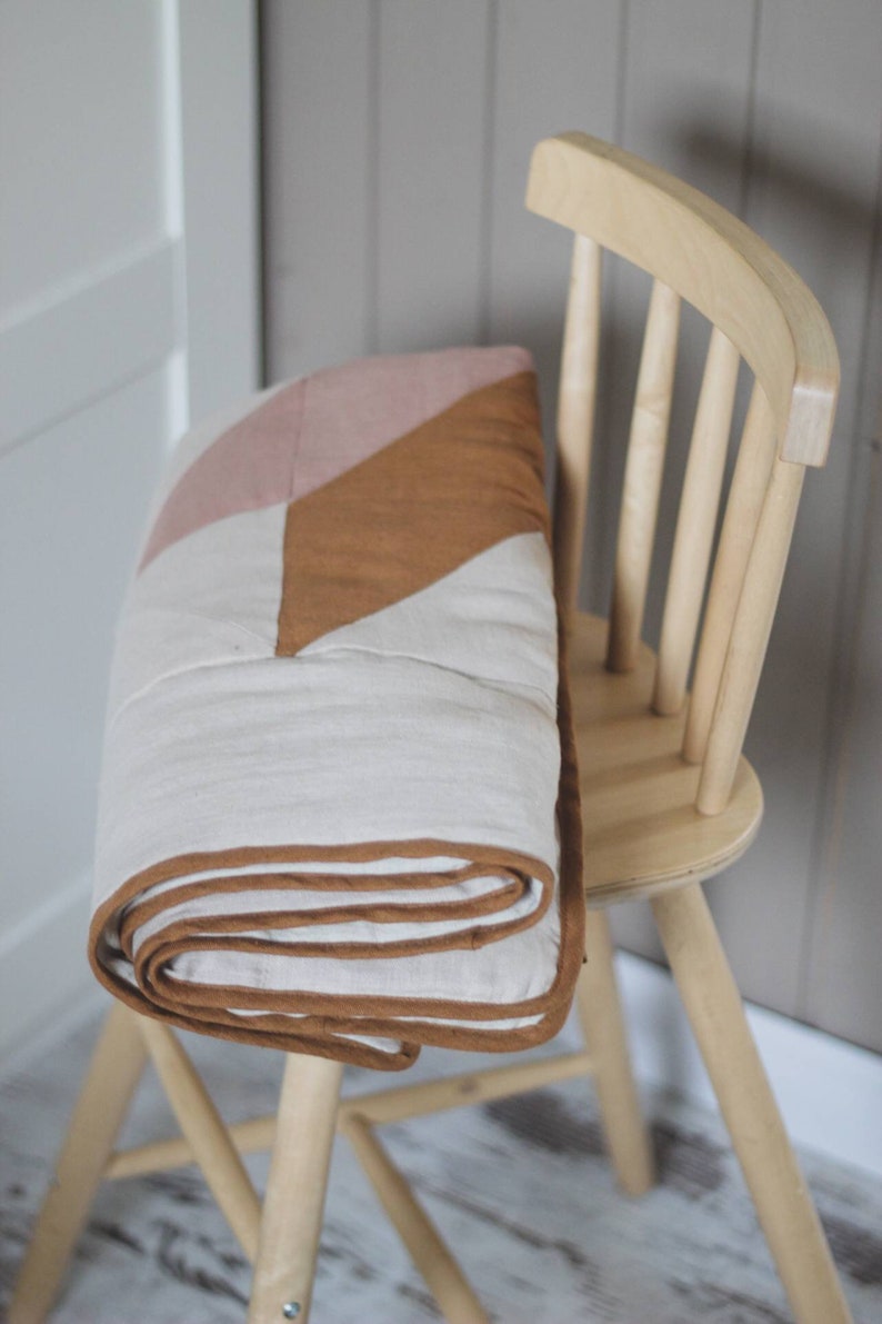 May include: A folded quilt with a geometric design in shades of white, pink, and brown rests on a light wooden chair. The quilt has a brown border and is neatly rolled. The chair is made of light-colored wood.