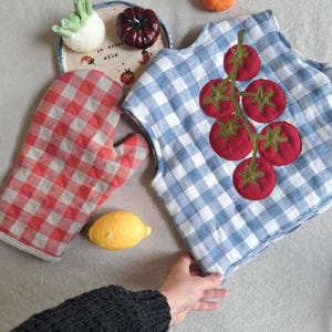 Puede incluir: Un guante de cocina a cuadros rojos y blancos y un delantal a cuadros azules y blancos con aplicaciones de tomate. También se ve un plato de cerámica con un diseño de tomate, un limón y una pequeña naranja.
