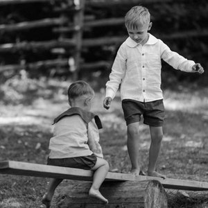 May include: Two young children playing on a seesaw made from a wooden plank and a log. One child is standing, wearing a white button-down shirt and dark shorts. The other child is seated, wearing a sailor-style top and shorts. The scene is in black and white.